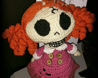 Puede incluir: Muñeca de ganchillo hecha a mano con una cara de calavera, ojos negros y pelo naranja. La muñeca lleva un vestido rosa con botones de madera y zapatos negros. Un artículo único hecho a mano.
