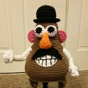 May include: A crocheted Mr. Potato Head doll with a black bowler hat, a black mustache, and an orange nose. The doll has white arms and hands, pink ears, and large, star-shaped eyes. The body is brown.