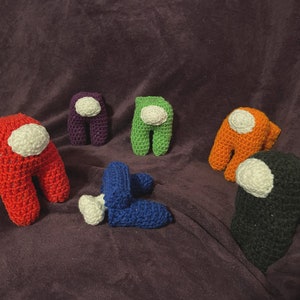 May include: A collection of six crocheted Among Us game characters in various colors: red, purple, green, orange, blue, and black. Each character has a white visor. The figures are arranged on a purple surface.