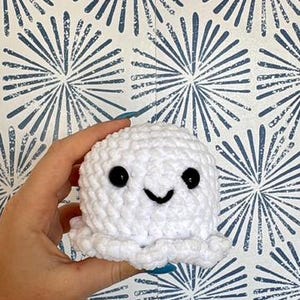 May include: A handmade, white crochet ghost with black eyes and a smiling mouth. The ghost is round with a textured surface. The background is a blue and white patterned wallpaper.