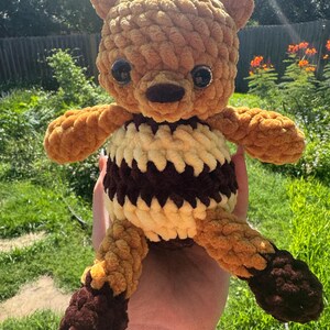 May include: A handmade, plush teddy bear in shades of brown, tan, and cream. The bear has a round head with small, dark eyes and a brown nose. Its body features a striped pattern of cream and brown, with brown paws and feet. The bear is made of a soft, textured yarn.