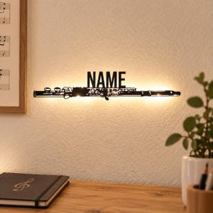 Peut inclure: Une décoration murale en métal noir en forme de flûte avec le mot NAME affiché en lettres capitales au-dessus de l'instrument. Le design intègre un rétroéclairage qui projette une lueur chaude sur le mur derrière la silhouette métallique.