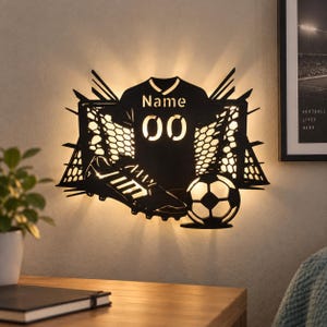 Pode incluir: Uma luminária de parede LED preta com a silhueta de uma camisola de futebol com o texto Name e 00, uma chuteira e uma bola de futebol. O design inclui um padrão de rede geométrico que emite uma luz quente na parede.
