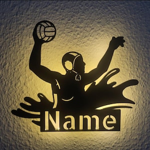 Può includere: Arte da parete in metallo nero raffigurante un giocatore di pallanuoto con una palla, che si protende verso l'alto. La silhouette include un design a spruzzo e una targhetta rettangolare con la parola "Name" ritagliata. L'arte è illuminata da dietro.