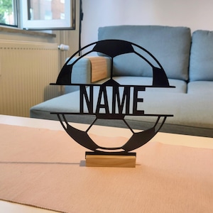 Puede incluir: Una silueta de bal&oacute;n de f&uacute;tbol negra, circular y cortada con l&aacute;ser, que presenta una barra horizontal en el centro con la palabra NAME en letras may&uacute;sculas y negritas. El objeto est&aacute; montado sobre una base rectangular de madera clara.