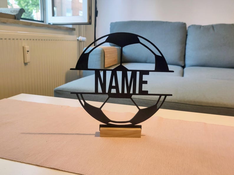 Puede incluir: Una silueta de bal&oacute;n de f&uacute;tbol negra, circular y cortada con l&aacute;ser, que presenta una barra horizontal en el centro con la palabra NAME en letras may&uacute;sculas y negritas. El objeto est&aacute; montado sobre una base rectangular de madera clara.
