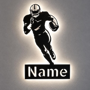 Pode incluir: Um letreiro de parede preto com a silhueta de um jogador de futebol americano em movimento segurando uma bola, posicionado acima de uma placa retangular com a palavra Name recortada, iluminado por uma luz de fundo branca suave.
