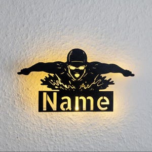 May include: Black metal wall art depicting a swimmer in the butterfly stroke. The swimmer wears goggles and a cap. Below the swimmer is a rectangular sign with the word "Name" cut out. The art is illuminated from behind.