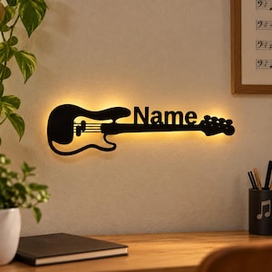 Könnte beinhalten: Ein schwarzes, wandmontiertes Schild in Form einer E-Gitarren-Silhouette, bei dem das Wort Name in den Hals integriert ist. Das ausgeschnittene Design wird von einem warmen gelben Licht hinterleuchtet, das einen sanften Halo-Effekt erzeugt.