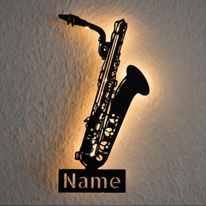 Könnte beinhalten: Schwarze Metall-Saxophon-Wandkunst mit dem Wort "Name" am unteren Rand ausgeschnitten. Das Saxophon wird von hinten beleuchtet, wodurch ein warmer Schein entsteht. Ein dekoratives Stück für Musikliebhaber.