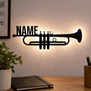 May include: A black metal wall-mounted light fixture shaped like a trumpet with the word NAME integrated into the design. The silhouette features a backlit glow that illuminates the wall behind the instrument-shaped frame, creating a soft halo effect.