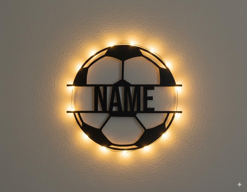 Puede incluir: Una decoraci&oacute;n de pared circular negra con forma de bal&oacute;n de f&uacute;tbol que presenta una barra horizontal en el centro con la palabra NAME en letras may&uacute;sculas. La pieza est&aacute; iluminada por luces LED de color blanco c&aacute;lido alrededor del marco.