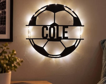 May include: A circular black metal wall sign shaped like a soccer ball with the name COLE cut out in the center. The sign is backlit with warm white LED lights that illuminate the wall behind the structure, creating a glowing effect.