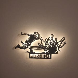 May include: A black metal wall art piece featuring a silhouette of a person bowling, with a ball striking pins and a rectangular base plate below containing the text WUNSCHTEXT. The design is backlit, creating a glowing effect around the edges.