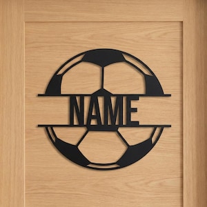 Puede incluir: Un letrero de pared circular de metal negro cortado con l&aacute;ser que presenta un dise&ntilde;o de bal&oacute;n de f&uacute;tbol dividido horizontalmente. La palabra NAME se muestra en letras may&uacute;sculas negritas y sin remates dentro del espacio central.