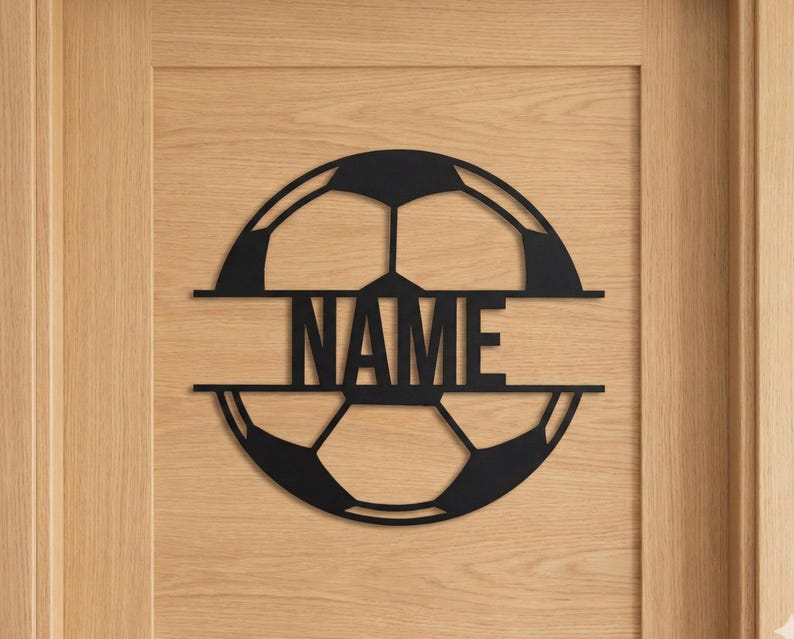 Puede incluir: Un letrero de pared circular de metal negro cortado con l&aacute;ser que presenta un dise&ntilde;o de bal&oacute;n de f&uacute;tbol dividido horizontalmente. La palabra NAME se muestra en letras may&uacute;sculas negritas y sin remates dentro del espacio central.