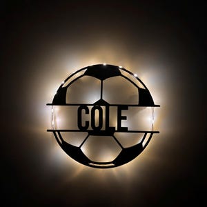 Puede incluir: Un letrero de pared circular de metal negro con forma de bal&oacute;n de f&uacute;tbol con una barra horizontal en el centro que presenta el nombre COLE en letras may&uacute;sculas negritas. El letrero est&aacute; retroiluminado con luces LED blancas c&aacute;lidas.