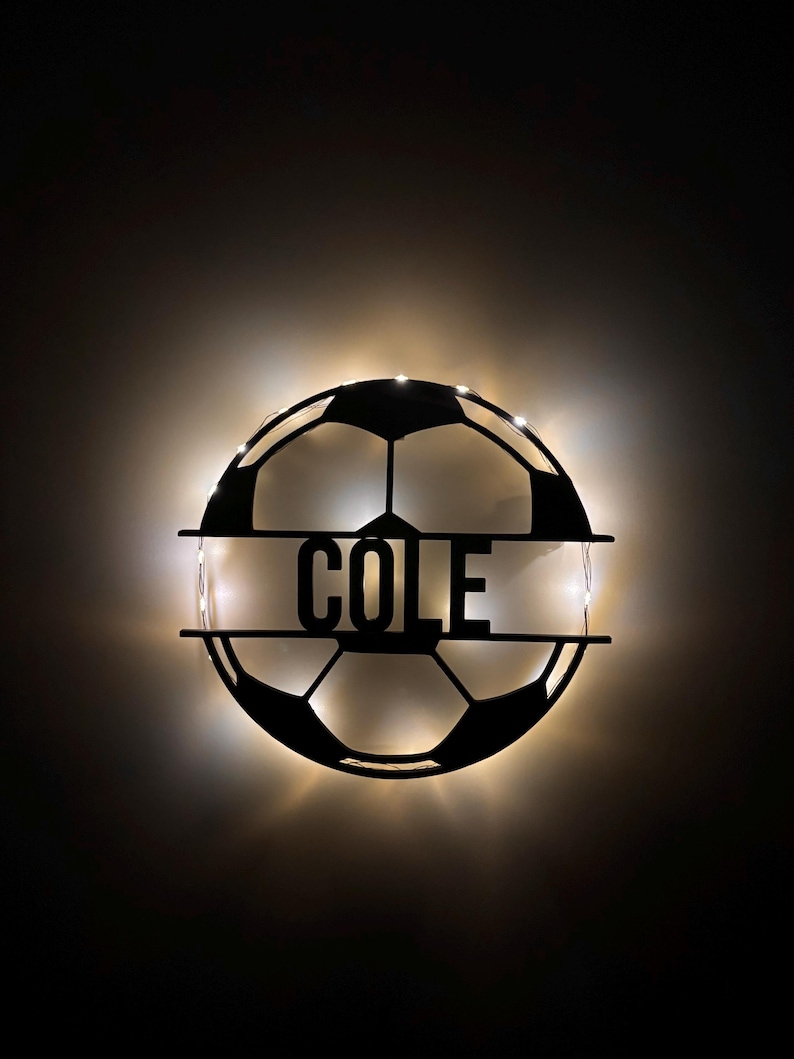Puede incluir: Un letrero de pared circular de metal negro con forma de bal&oacute;n de f&uacute;tbol con una barra horizontal en el centro que presenta el nombre COLE en letras may&uacute;sculas negritas. El letrero est&aacute; retroiluminado con luces LED blancas c&aacute;lidas.