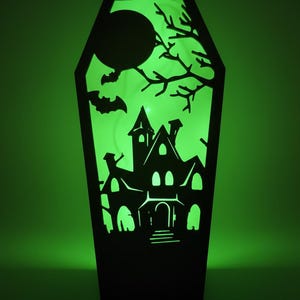 May include: A black coffin-shaped Halloween decoration featuring a haunted house silhouette, bats, a full moon, and bare tree branches. The interior glows with green light, creating a spooky atmosphere. The decoration is made of wood or a similar material.