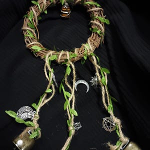 Może przedstawiać: Rustykalna, ręcznie wykonana dekoracja ścienna z plecionym wieńcem z winorośli ozdobionym sztucznymi zielonymi liśćmi i sznurkiem. Z wieńca zwisają trzy pasma z metalowymi zawieszkami, sztucznymi liśćmi i małymi dzwoneczkami. Brązowy i pomarańczowy kamień zawieszony jest na górze.