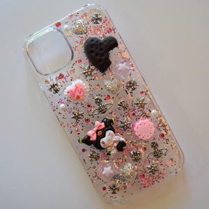 Puede incluir: Funda transparente para tel&eacute;fono decorada con purpurina, estrellas, corazones y adornos en forma de flor. Presenta una galleta negra en forma de coraz&oacute;n con la palabra "TORUN" y un personaje de dibujos animados con un lazo rosa.