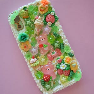 Puede incluir: Funda de teléfono decorativa con un diseño caprichoso. Presenta ositos de goma rosas, fresas, conos de helado y detalles florales. Tiene un borde blanco texturizado y una combinación de colores verde y blanco.