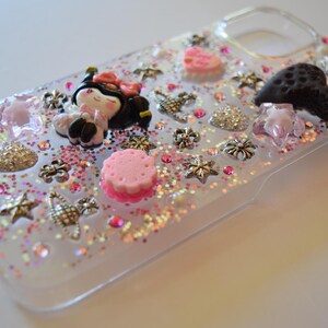 Puede incluir: Funda de tel&eacute;fono transparente decorada con purpurina rosa, una peque&ntilde;a mu&ntilde;eca y varios abalorios. Los abalorios incluyen estrellas, flores y un coraz&oacute;n. La funda tiene un borde transparente y est&aacute; llena de purpurina rosa y plateada.