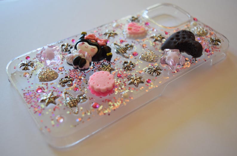 Puede incluir: Funda de tel&eacute;fono transparente decorada con purpurina rosa, una peque&ntilde;a mu&ntilde;eca y varios abalorios. Los abalorios incluyen estrellas, flores y un coraz&oacute;n. La funda tiene un borde transparente y est&aacute; llena de purpurina rosa y plateada.