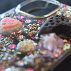 Puede incluir: Una funda transparente para tel&eacute;fono decorada con varios abalorios y adornos. La funda presenta un abalorio en forma de coraz&oacute;n rosa con el texto "Birthday Day", un abalorio en forma de coraz&oacute;n marr&oacute;n oscuro y varios abalorios en forma de estrella. La funda tambi&eacute;n est&aacute; adornada con purpurina y pedrer&iacute;a.