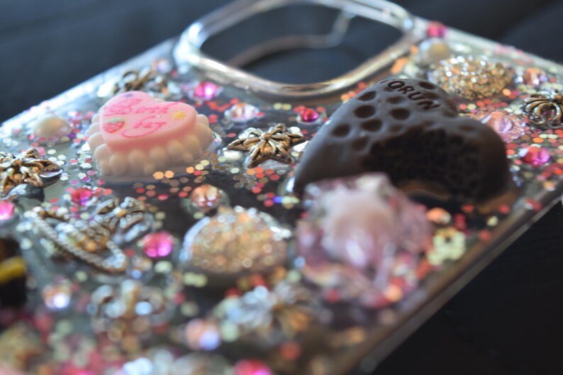Puede incluir: Una funda transparente para tel&eacute;fono decorada con varios abalorios y adornos. La funda presenta un abalorio en forma de coraz&oacute;n rosa con el texto "Birthday Day", un abalorio en forma de coraz&oacute;n marr&oacute;n oscuro y varios abalorios en forma de estrella. La funda tambi&eacute;n est&aacute; adornada con purpurina y pedrer&iacute;a.