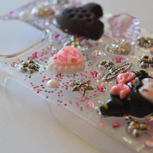Puede incluir: Funda de tel&eacute;fono transparente decorada con varios abalorios, incluyendo un coraz&oacute;n negro, un pastel rosa en forma de coraz&oacute;n y una figura de Minnie Mouse. La funda est&aacute; adornada con purpurina rosa y peque&ntilde;as estrellas decorativas.