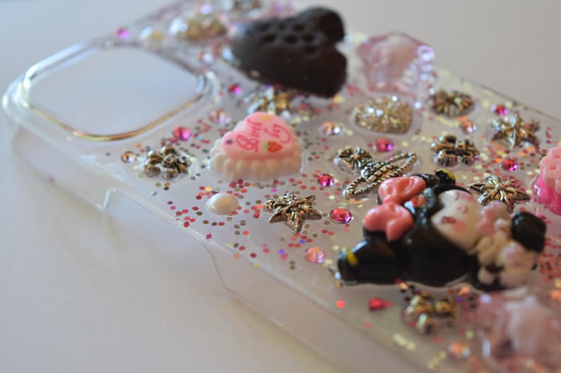 Puede incluir: Funda de tel&eacute;fono transparente decorada con varios abalorios, incluyendo un coraz&oacute;n negro, un pastel rosa en forma de coraz&oacute;n y una figura de Minnie Mouse. La funda est&aacute; adornada con purpurina rosa y peque&ntilde;as estrellas decorativas.