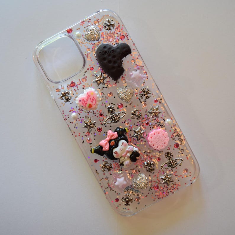 Puede incluir: Funda transparente para tel&eacute;fono decorada con purpurina, estrellas, corazones y adornos en forma de flor. Presenta una galleta negra en forma de coraz&oacute;n con la palabra "TORUN" y un personaje de dibujos animados con un lazo rosa.