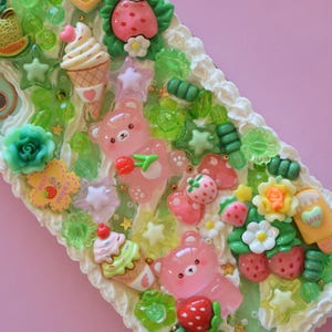 Puede incluir: Un objeto rectangular decorativo con fondo rosa, con un dise&ntilde;o caprichoso de osos rosas, fresas, conos de helado y detalles en forma de estrella. Los bordes est&aacute;n adornados con glaseado blanco.