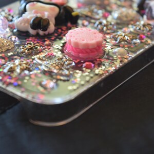 Puede incluir: Una funda transparente para tel&eacute;fono decorada con purpurina rosa, dorada y plateada, junto con varios adornos. La funda presenta un dije de pastel rosa en capas, una figura blanca y negra y varios dijes decorativos.