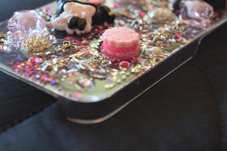 Puede incluir: Una funda transparente para tel&eacute;fono decorada con purpurina rosa, dorada y plateada, junto con varios adornos. La funda presenta un dije de pastel rosa en capas, una figura blanca y negra y varios dijes decorativos.
