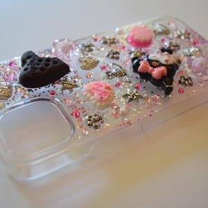 Puede incluir: Funda de tel&eacute;fono transparente decorada con purpurina rosa y dorada, varios abalorios y pedrer&iacute;a. Los abalorios incluyen un zapato negro, un pastel rosa y un gato negro con un lazo rosa. La funda tiene un recorte para la c&aacute;mara y los botones.