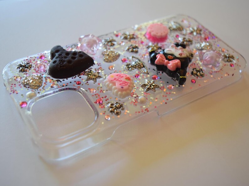 Puede incluir: Funda de tel&eacute;fono transparente decorada con purpurina rosa y dorada, varios abalorios y pedrer&iacute;a. Los abalorios incluyen un zapato negro, un pastel rosa y un gato negro con un lazo rosa. La funda tiene un recorte para la c&aacute;mara y los botones.