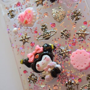 Puede incluir: Funda de tel&eacute;fono transparente decorada con varios abalorios, incluyendo un coraz&oacute;n, estrellas y un personaje de dibujos animados con un lazo rosa. La funda est&aacute; cubierta de purpurina rosa e iridiscente y peque&ntilde;os brillantes rosas.