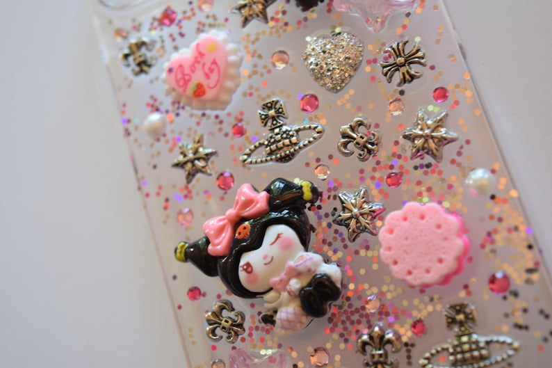 Puede incluir: Funda de tel&eacute;fono transparente decorada con varios abalorios, incluyendo un coraz&oacute;n, estrellas y un personaje de dibujos animados con un lazo rosa. La funda est&aacute; cubierta de purpurina rosa e iridiscente y peque&ntilde;os brillantes rosas.