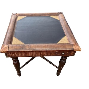 May include: Square, antique-style table with a dark leather top and a woven, textured border. The table has dark brown, turned legs and a lower shelf. The corners are reinforced with metal. A stylish piece of furniture.