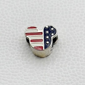 Puede incluir: Un charm de plata con forma de cabeza de Mickey Mouse, con un dise&ntilde;o patri&oacute;tico. El lado izquierdo tiene rayas rojas y blancas, mientras que el lado derecho tiene un fondo azul con estrellas blancas, similar a la bandera estadounidense. El charm est&aacute; sobre una superficie blanca texturizada.