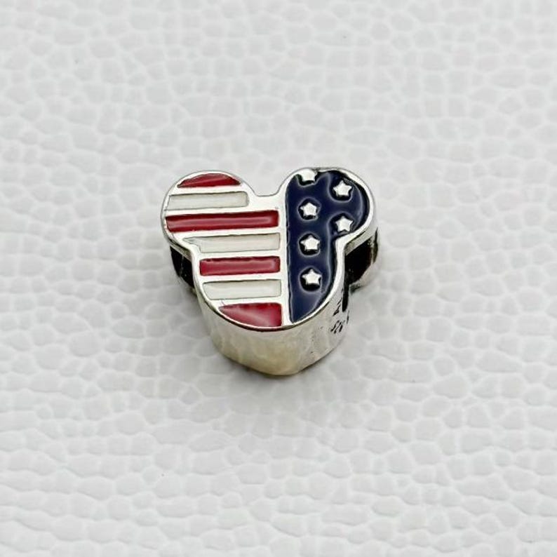 Puede incluir: Un charm de plata con forma de cabeza de Mickey Mouse, con un dise&ntilde;o patri&oacute;tico. El lado izquierdo tiene rayas rojas y blancas, mientras que el lado derecho tiene un fondo azul con estrellas blancas, similar a la bandera estadounidense. El charm est&aacute; sobre una superficie blanca texturizada.