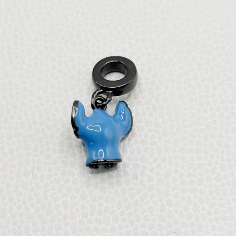 Puede incluir: Un charm azul y negro con un dise&ntilde;o de personaje de dibujos animados. El charm tiene un cuerpo azul claro con detalles en negro y un anillo negro en la parte superior para sujetarlo. El charm es probablemente para una pulsera o collar.
