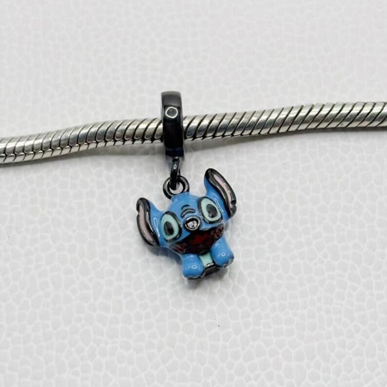 Puede incluir: Un charm de esmalte azul con el personaje Stitch de Lilo & Stitch. El charm tiene una anilla negra y est&aacute; unido a una cadena plateada. Stitch tiene orejas grandes, boca roja y cuerpo azul claro.