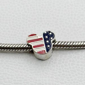 Puede incluir: Un charm de plata con forma de cabeza de rat&oacute;n, con un dise&ntilde;o patri&oacute;tico. El charm est&aacute; dividido en secciones con rayas rojas y blancas y un &aacute;rea azul con estrellas blancas, que se asemeja a la bandera estadounidense. Est&aacute; ensartado en una pulsera de plata.