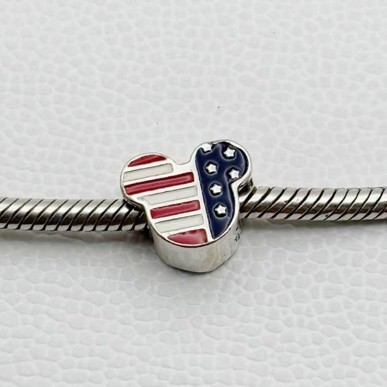 Puede incluir: Un charm de plata con forma de cabeza de rat&oacute;n, con un dise&ntilde;o patri&oacute;tico. El charm est&aacute; dividido en secciones con rayas rojas y blancas y un &aacute;rea azul con estrellas blancas, que se asemeja a la bandera estadounidense. Est&aacute; ensartado en una pulsera de plata.