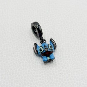 Puede incluir: Un charm azul y negro con un personaje de dibujos animados con orejas grandes y boca roja. El charm tiene un lazo negro para sujetarlo a una pulsera o collar. La cara del personaje est&aacute; detallada con detalles blancos y negros.