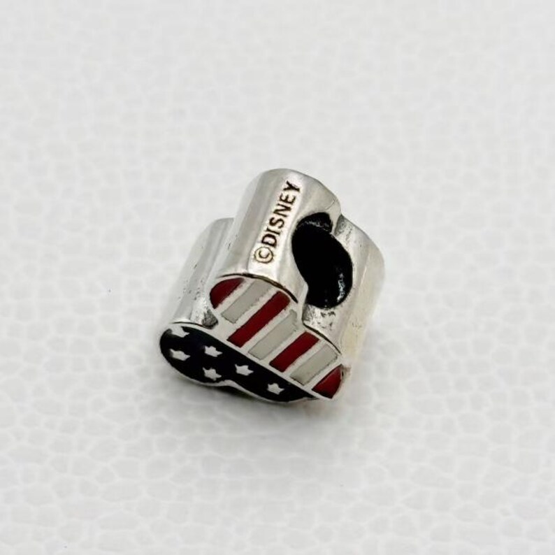 Puede incluir: Un charm plateado con un dise&ntilde;o patri&oacute;tico. El charm presenta rayas rojas y blancas, estrellas azules y el texto "&copy;DISNEY". Este charm es probablemente un objeto de colecci&oacute;n o una joya.