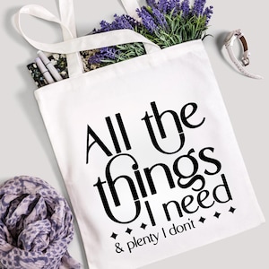 K&ouml;nnte beinhalten: Wei&szlig;e Tragetasche mit dem Text "All the things I need & plenty I don't" in Schwarz. Die Tasche enth&auml;lt Lavendel, Stifte und ein Blumennotizbuch. Ein Schal und eine Uhr liegen neben der Tasche.